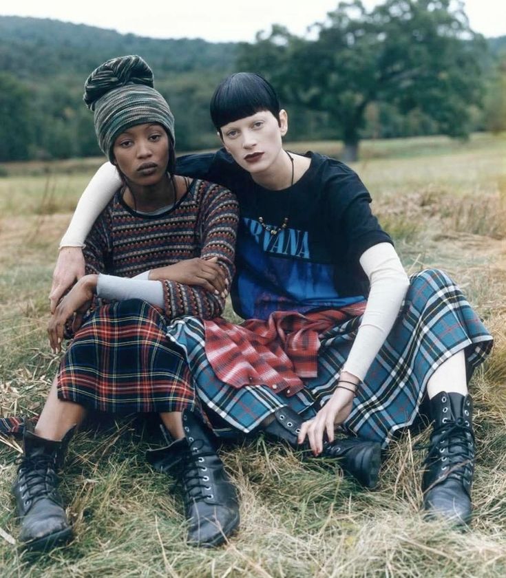 Two individuals in stylish plaid outfits sit outdoors, expressing friendship and alternative fashion in a serene natural setting.