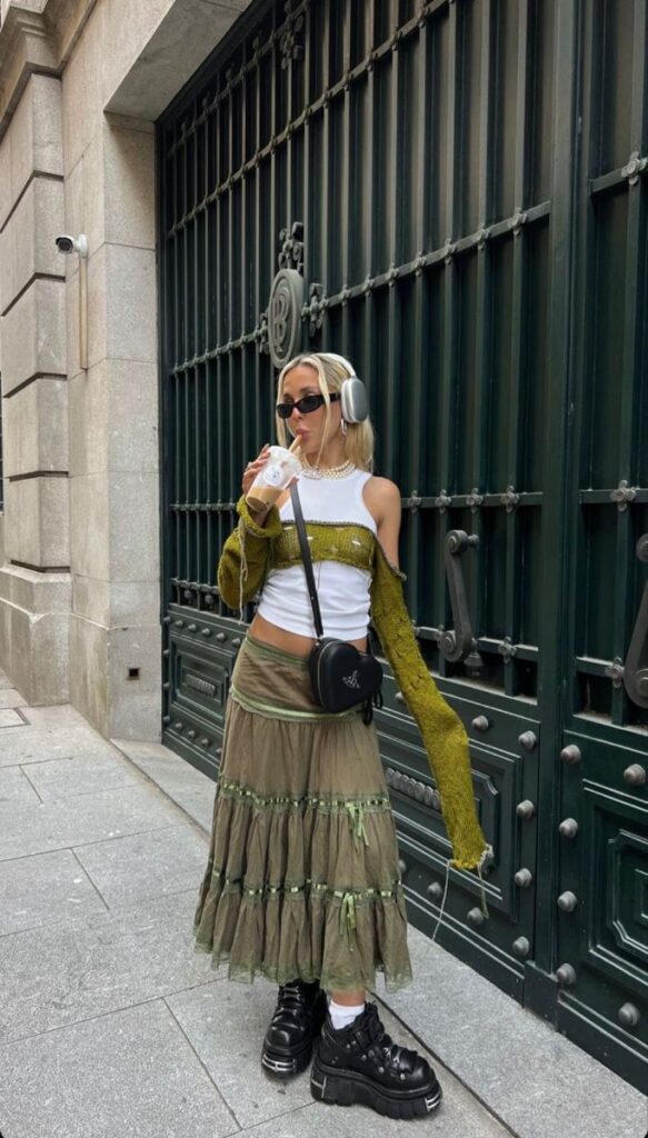Trendsetting fashionista in green skirt, crop top, and headphones drinks iced coffee, leaning against a stylish gate.