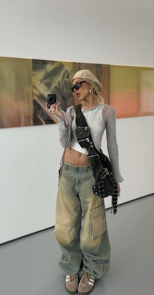 Fashionable person in a chic, artistic outfit with a beige hat, textured top, and cargo pants taking a mirror selfie in a gallery.
