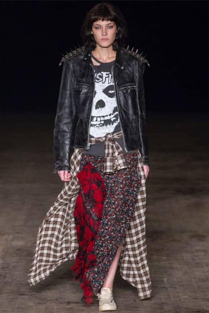 Model in punk fashion: spiked leather jacket, skull tee, plaid and floral skirt on runway. Style mix of grunge, punk, and vintage elements.