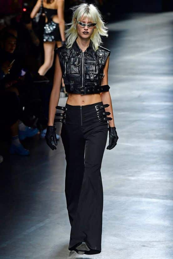 Model walks runway in edgy black leather outfit with crop vest and flared pants at fashion show, wearing sunglasses and gloves.