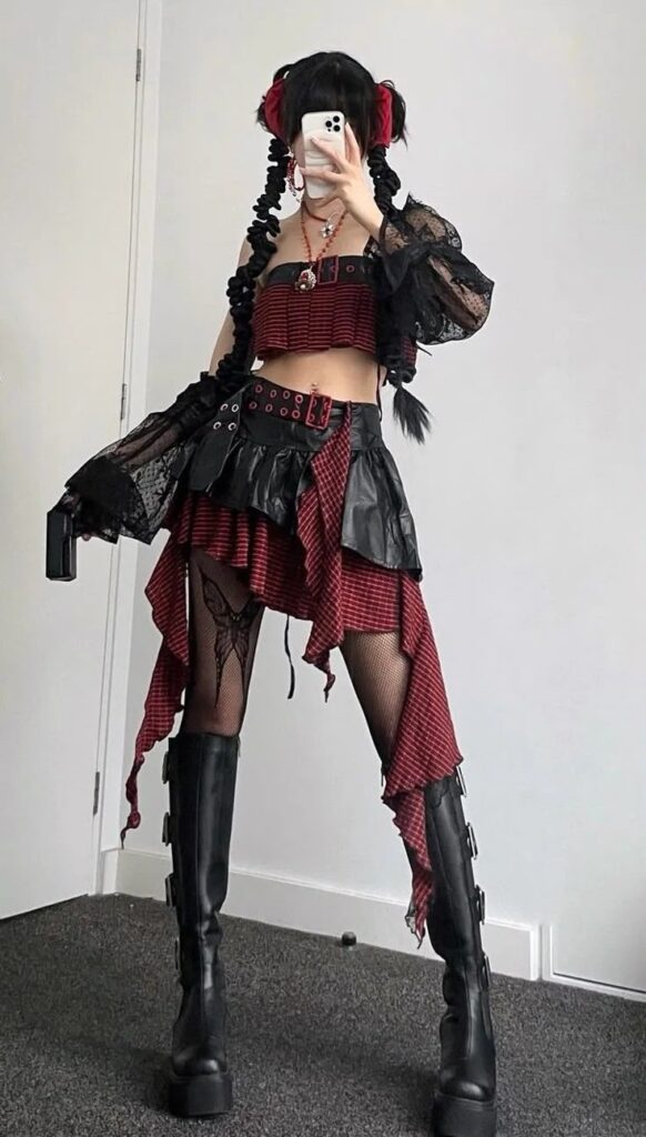Person in edgy goth outfit with red plaid skirt, lace sleeves, and tall black boots, taking a mirror selfie.