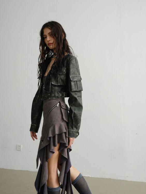 Fashion model in edgy outfit with leather jacket and asymmetrical skirt standing in minimalistic room.