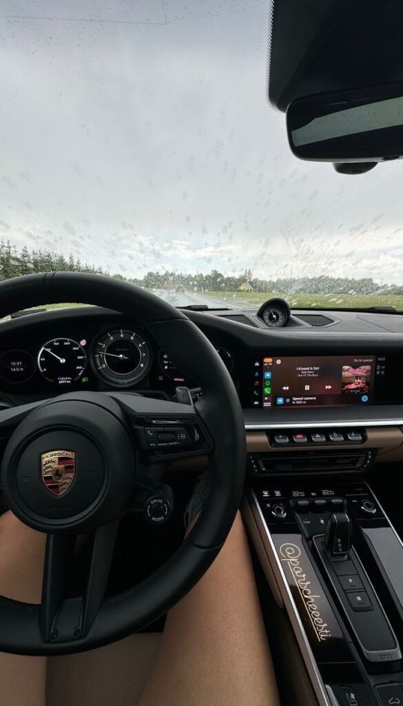 Inside view of a luxury car cockpit with music display, dashboard controls, and rainy windshield through the steering wheel.