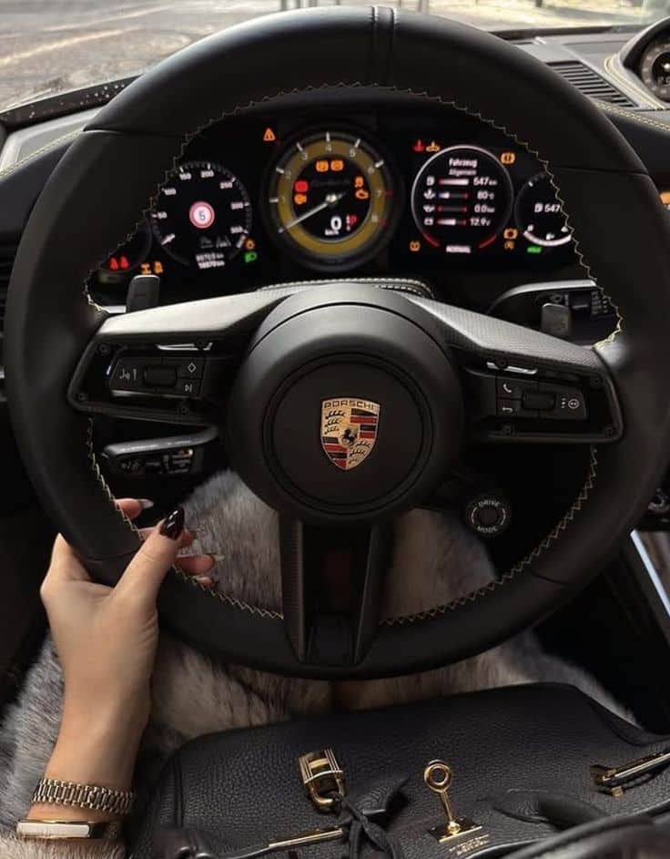 Close-up of a hand on a luxury car steering wheel, dashboard displaying gauges, and a designer bag in view.
