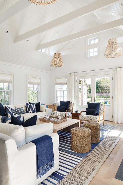 Bright coastal living room with white walls, blue accents, and wicker decor. Features large windows, ceiling beams, and natural light.
