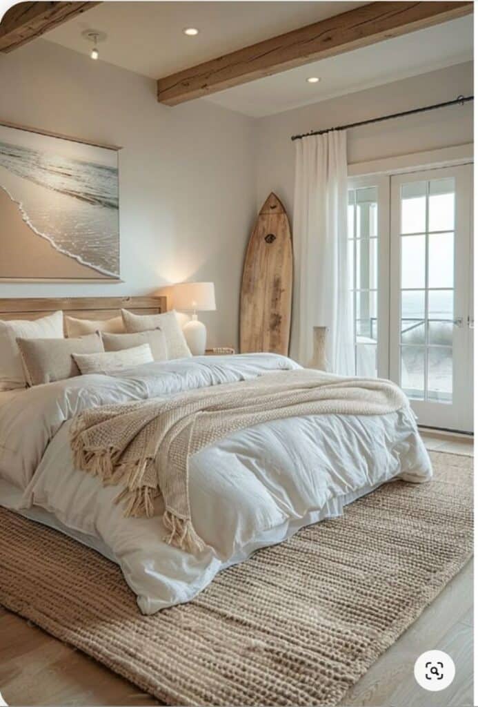 Beach-themed bedroom with cozy white bedding, rustic surfboard decor, and natural light from a window near the ocean.
