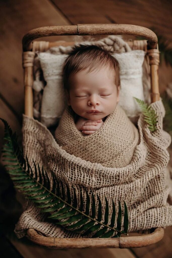 _ - 2025-12-12T130231690 Newborn baby peacefully sleeping in a cozy wicker basket, wrapped in a soft, brown blanket with a fern leaf accent. | Sky Rye Design Newborn baby peacefully sleeping in a cozy wicker basket, wrapped in a soft, brown blanket with a fern leaf accent.