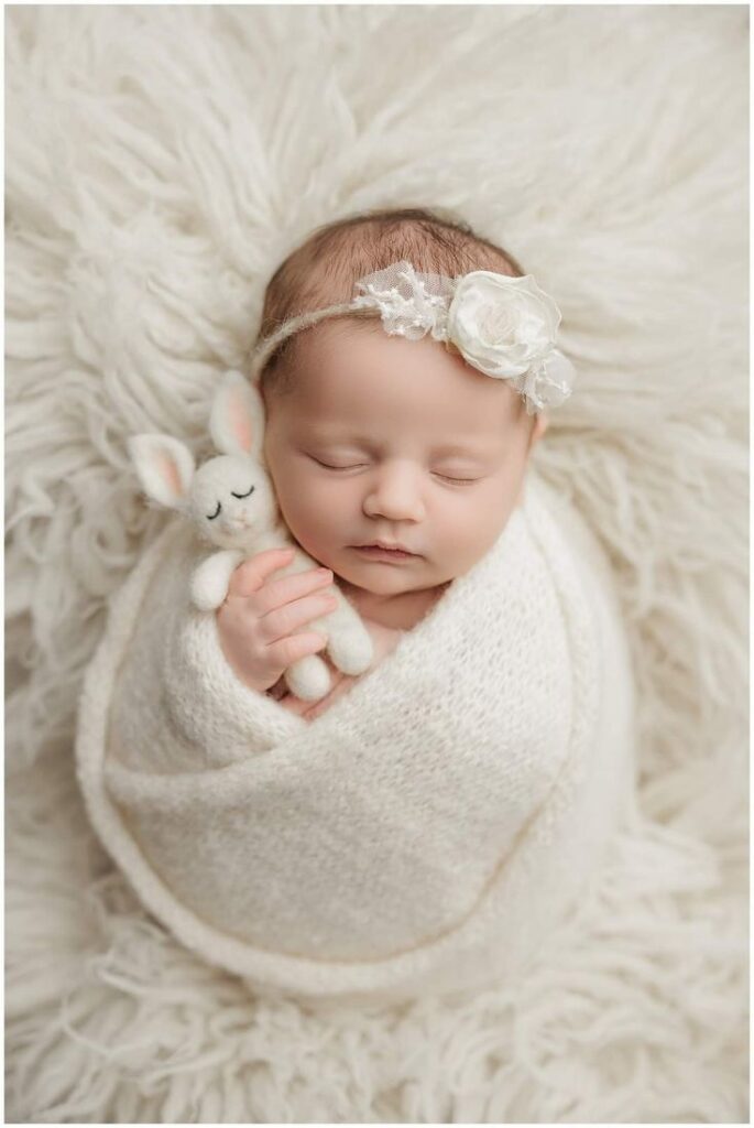 _ - 2025-12-12T131634497 Sleeping baby in white wrap, holding a small plush bunny, wearing a floral headband on a soft, fluffy blanket. | Sky Rye Design Sleeping baby in white wrap, holding a small plush bunny, wearing a floral headband on a soft, fluffy blanket.