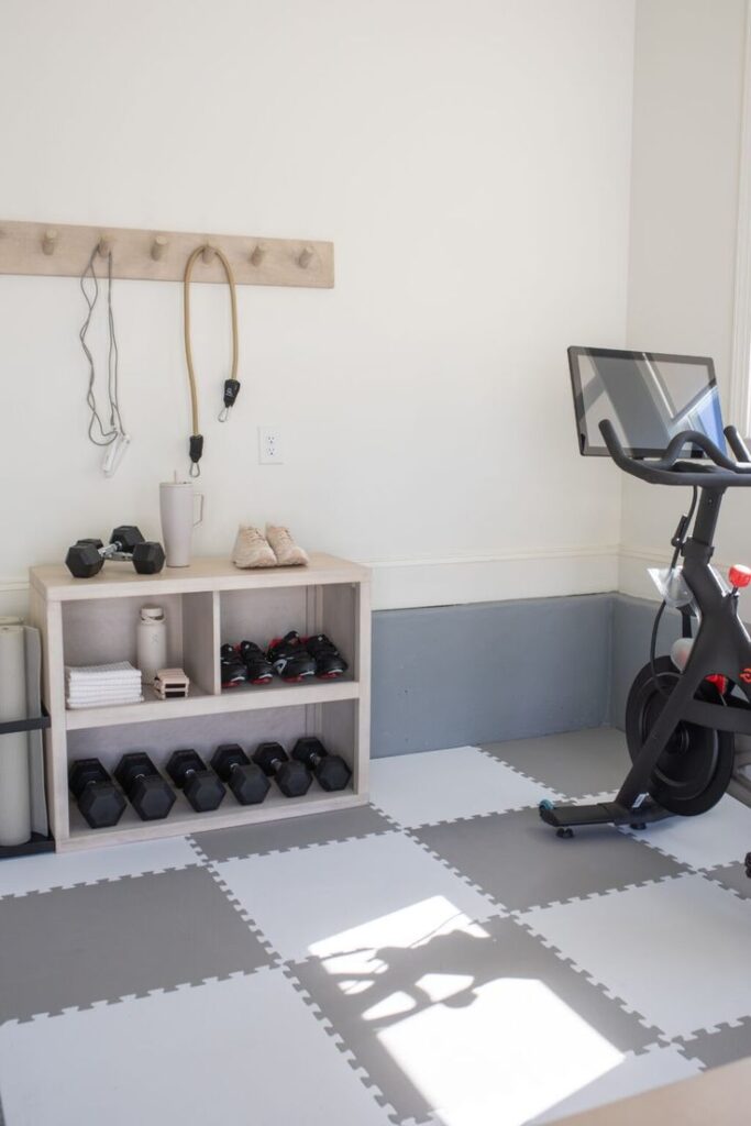 _ - 2025-12-16T232156368 Home gym setup with exercise bike, dumbbells, and storage shelves on padded flooring, promoting a functional and organized fitness area. | Sky Rye Design Home gym setup with exercise bike, dumbbells, and storage shelves on padded flooring, promoting a functional and organized fitness area.