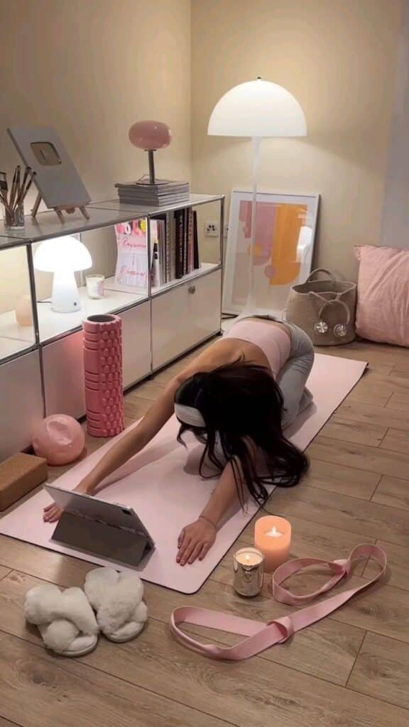 _ - 2025-12-16T232206708 Woman practicing yoga at home on a pink mat with candles and a tablet, surrounded by cozy decor and exercise equipment. | Sky Rye Design Woman practicing yoga at home on a pink mat with candles and a tablet, surrounded by cozy decor and exercise equipment.