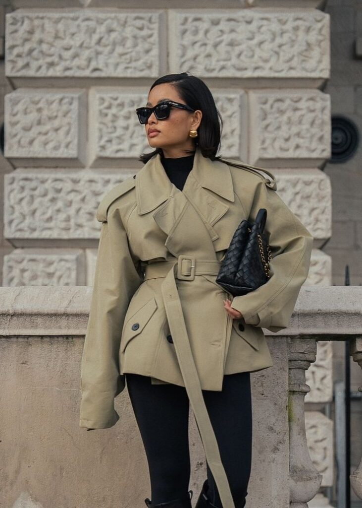 _ - 2025-12-28T121753027 Stylish woman in oversized trench coat and sunglasses, standing confidently against a textured wall, holding a chic black handbag. | Sky Rye Design Stylish woman in oversized trench coat and sunglasses, standing confidently against a textured wall, holding a chic black handbag.