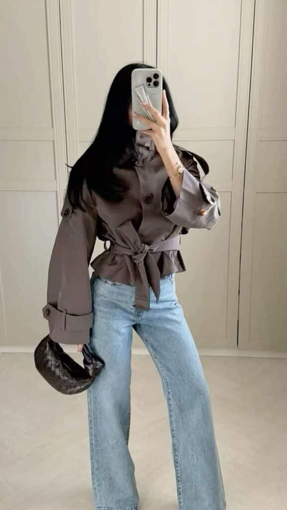 _ - 2025-12-28T122105499 Woman in stylish trench coat and jeans, taking mirror selfie. Holding small handbag and smartphone. Fashionable casual outfit. | Sky Rye Design Woman in stylish trench coat and jeans, taking mirror selfie. Holding small handbag and smartphone. Fashionable casual outfit.