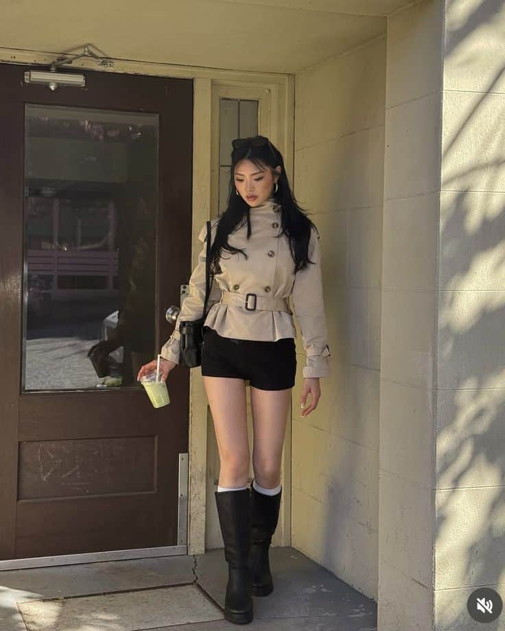 _ - 2025-12-28T122235192 Stylish woman in a beige trench coat, black shorts, and boots, holding a green drink, walking through a doorway. Cozy urban fashion. | Sky Rye Design Stylish woman in a beige trench coat, black shorts, and boots, holding a green drink, walking through a doorway. Cozy urban fashion.