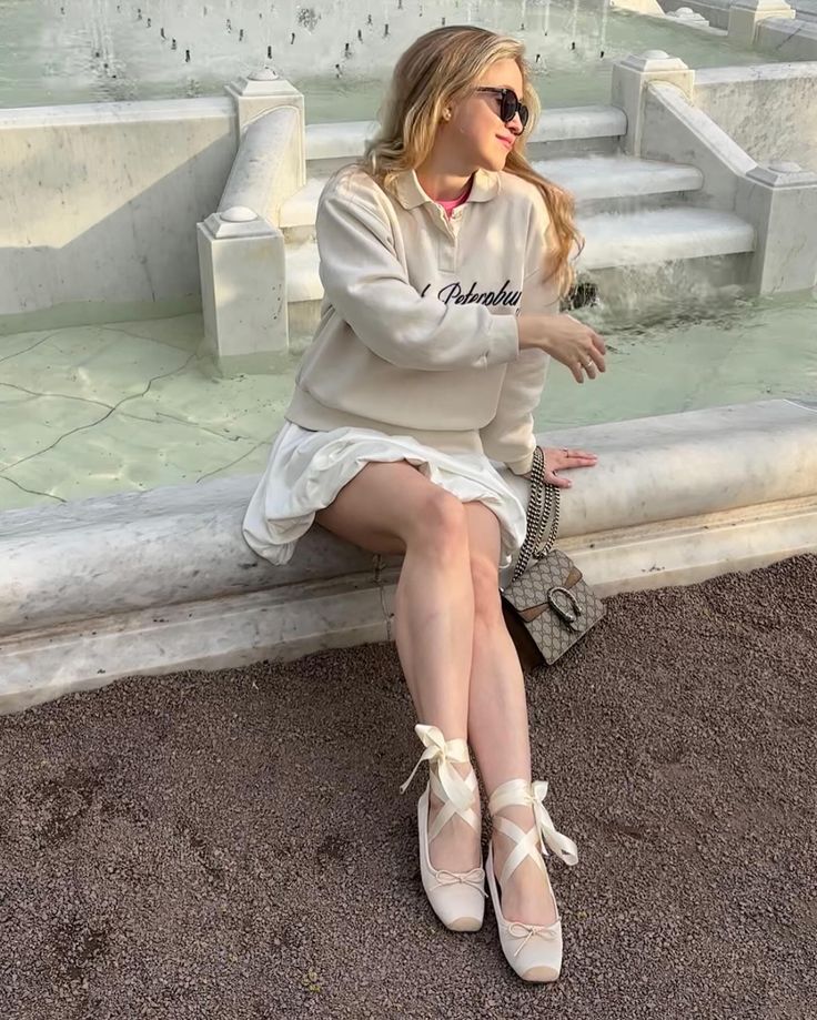 Woman in a stylish outfit sitting on a fountain ledge, wearing ballet flats and sunglasses, enjoying a sunny day outdoors.