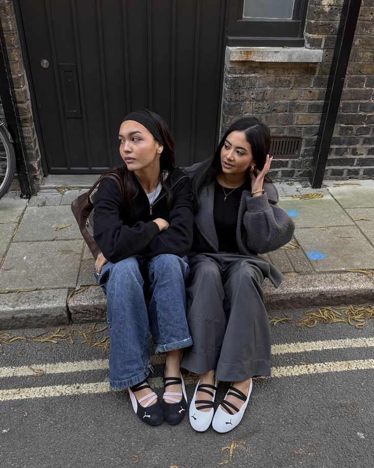 _ - 2025-12-28T125121617 Two women sitting on a curbside, wearing casual outfits and ballet flats, sharing a moment outdoors with a brick wall backdrop. | Sky Rye Design Two women sitting on a curbside, wearing casual outfits and ballet flats, sharing a moment outdoors with a brick wall backdrop.
