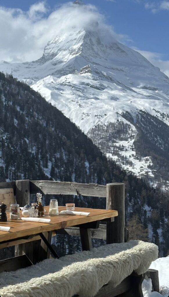 Screenshot Outdoor dining with stunning snowy mountain view, set table prepared for guests, cozy wooden bench with fur seating, sunlight shining. | Sky Rye Design Outdoor dining with stunning snowy mountain view, set table prepared for guests, cozy wooden bench with fur seating, sunlight shining.