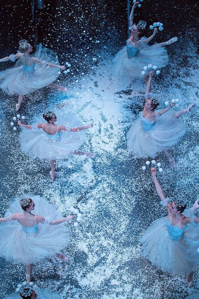 Ballet dancers in blue tutus perform gracefully amidst falling snow on stage, creating a magical winter scene.
