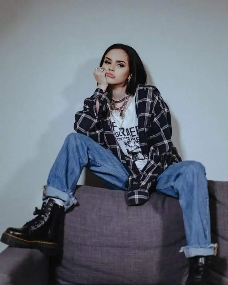 Fashionable person sitting on a couch in casual wear, plaid shirt, jeans, and boots, exuding a confident and edgy style.