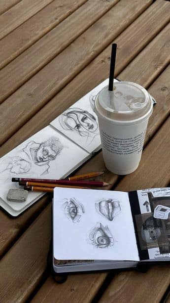 Open sketchbooks with pencil drawings, pencils, an eraser, and a coffee cup with a straw on a wooden table. Artistic workspace setup.