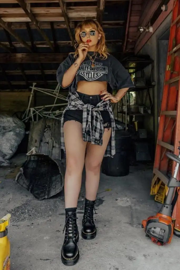 Trendy woman in edgy fashion, wearing a Harley-Davidson tee, plaid shirt, shorts, and chunky boots, posing in a rustic garage setting.