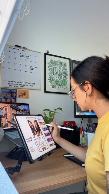 Woman using a stylus on a tablet in a home office, with a calendar and art on the wall, embodying creativity and productivity.