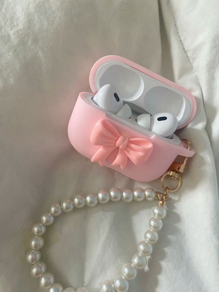 _ 24 Pink AirPods case with bow and pearl strap, placed on a white fabric surface. Stylish accessory for wireless earbuds. | Sky Rye Design Pink AirPods case with bow and pearl strap, placed on a white fabric surface. Stylish accessory for wireless earbuds.