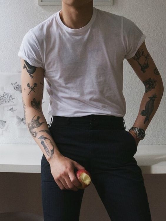 Person in white shirt and black pants showcasing tattoos, holding an apple. Stylish and modern casual attire with artistic arm designs.