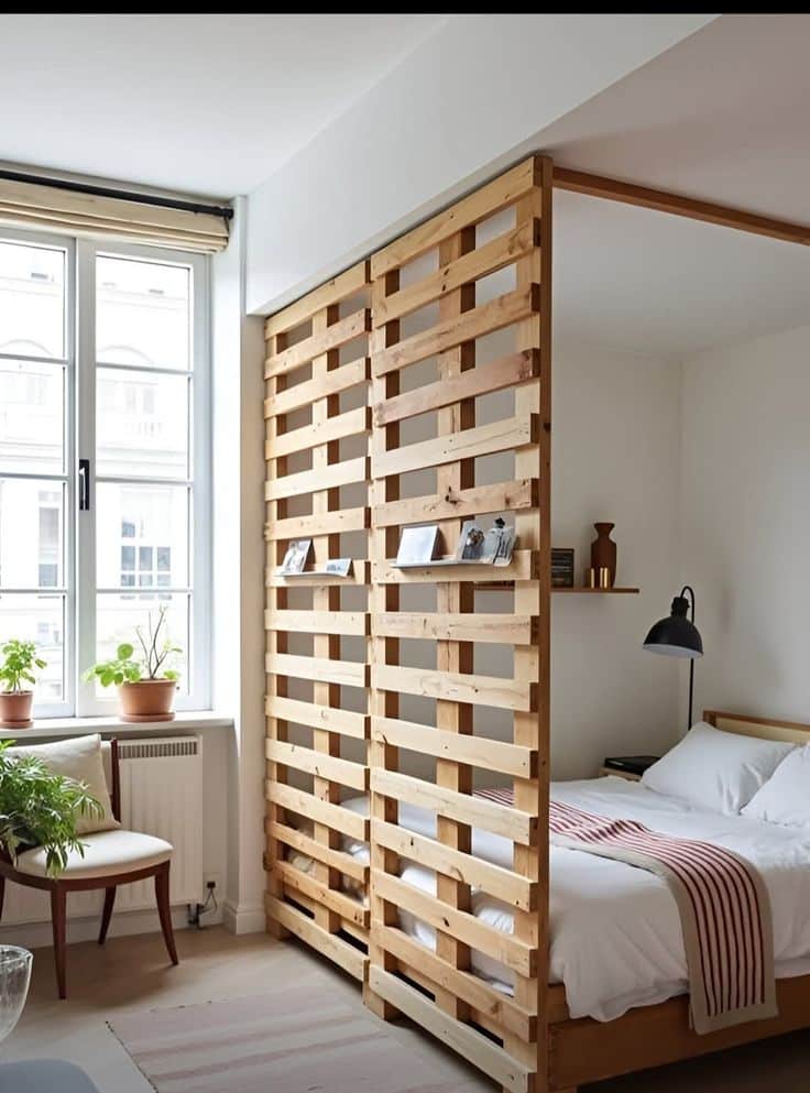_ 25 Cozy bedroom with wood pallet divider, bright window, and indoor plants. Modern design with striped blanket and wall lamp. | Sky Rye Design Cozy bedroom with wood pallet divider, bright window, and indoor plants. Modern design with striped blanket and wall lamp.