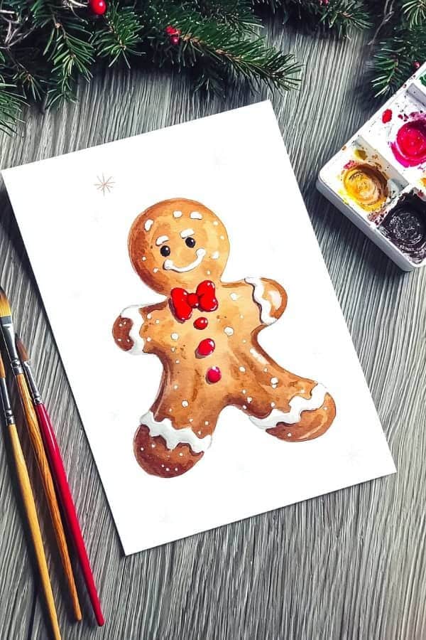 25 Watercolor Christmas Cards Ideas for DIY Holiday Inspiration 1 Watercolor painting of a smiling gingerbread man with a red bowtie, surrounded by paintbrushes and holiday greenery. | Sky Rye Design Watercolor painting of a smiling gingerbread man with a red bowtie, surrounded by paintbrushes and holiday greenery.