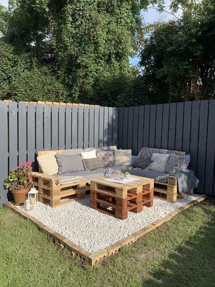 28909526b4d5f854ae6831d7fcd9b7b1 Cozy outdoor seating area with pallet furniture, cushions, and a small table on a gravel patio, bordered by a wooden fence and greenery. | Sky Rye Design Cozy outdoor seating area with pallet furniture, cushions, and a small table on a gravel patio, bordered by a wooden fence and greenery.