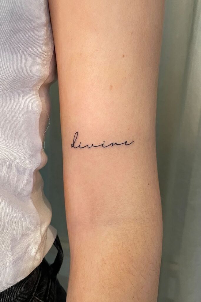 Simple divine script tattoo on inner arm in black ink, showcasing minimalist style and personal expression.
