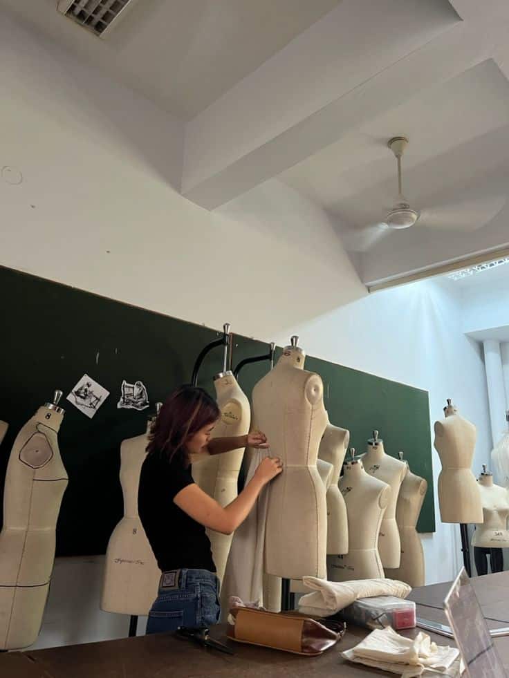 _ 3 Fashion design student fitting fabric on dress form in classroom workshop with multiple mannequins and sewing tools. | Sky Rye Design Fashion design student fitting fabric on dress form in classroom workshop with multiple mannequins and sewing tools.