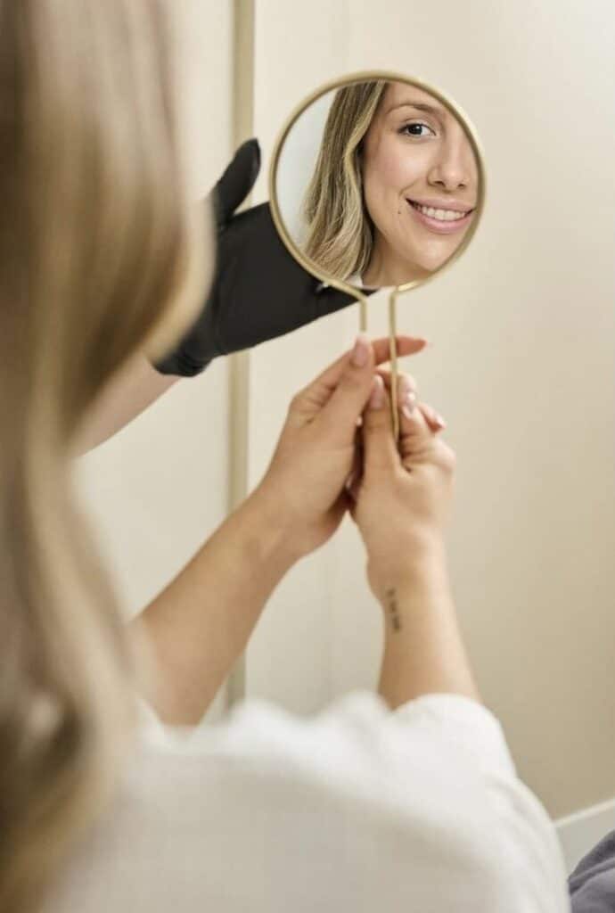 Screenshot Woman smiling in a round mirror held by gloved hand, illustrating self-reflection and positivity. | Sky Rye Design Woman smiling in a round mirror held by gloved hand, illustrating self-reflection and positivity.