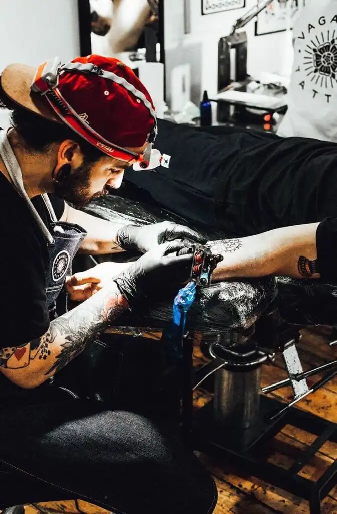 Tattoo artist in a red cap working on a detailed forearm design in a tattoo studio, using a tattoo machine and wearing gloves.