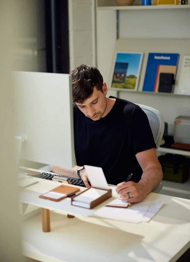 _ 3 Man in black shirt working at a desk, writing notes and using a computer in a modern office setting with shelves in the background. | Sky Rye Design Man in black shirt working at a desk, writing notes and using a computer in a modern office setting with shelves in the background.