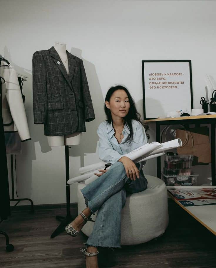 _ 3 Fashion designer sitting beside a mannequin with a plaid blazer, holding sketches in a stylish studio with sewing materials. | Sky Rye Design Fashion designer sitting beside a mannequin with a plaid blazer, holding sketches in a stylish studio with sewing materials.