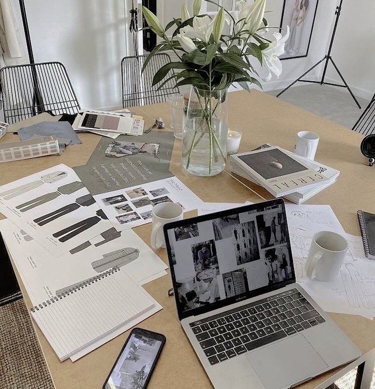 Design studio workspace with a laptop, sketches, magazines, and a vase of lilies on a table, conveying creativity and inspiration.