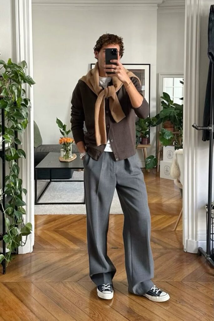 Man wearing casual cardigan and wide-leg trousers, taking mirror selfie in stylish living room with plants and hardwood floor.