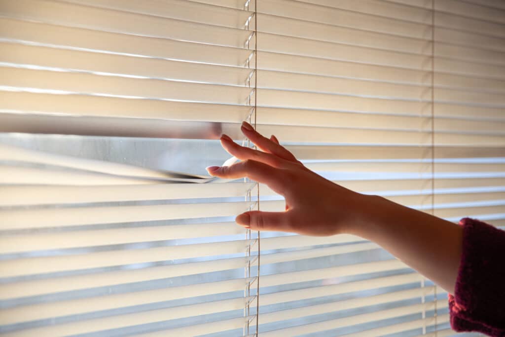 Womans hand taking a peak through the window blinds Hand adjusting horizontal blinds, letting in sunlight through a window, highlighting a cozy indoor atmosphere. | Sky Rye Design Hand adjusting horizontal blinds, letting in sunlight through a window, highlighting a cozy indoor atmosphere.