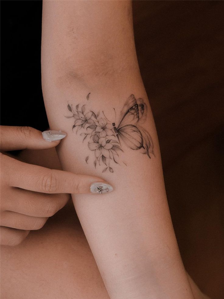 31 Beautiful Flower Tattoos for Women Delicate butterfly and floral tattoo on forearm, with person pointing, showcasing detailed ink art in soft lighting. | Sky Rye Design Delicate butterfly and floral tattoo on forearm, with person pointing, showcasing detailed ink art in soft lighting.