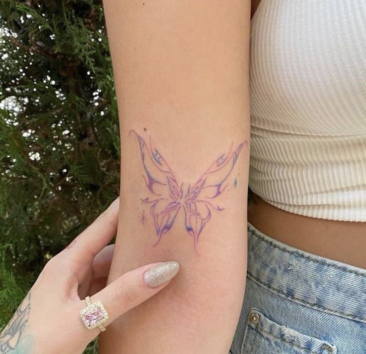 _ 35 Delicate butterfly tattoo in pastel shades on a woman's arm, wearing a white top and denim, with a hand showcasing it. | Sky Rye Design Delicate butterfly tattoo in pastel shades on a woman's arm, wearing a white top and denim, with a hand showcasing it.