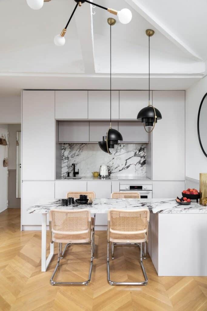 Modern kitchen with marble countertops, black pendant lights, and wicker chairs on a herringbone floor. Sleek, minimalist design.
