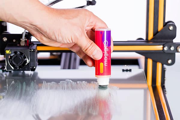 3D-Printer-Glue-Stick Hand applying glue stick to 3D printer bed for better adhesion, with printer visible in background. | Sky Rye Design Hand applying glue stick to 3D printer bed for better adhesion, with printer visible in background.