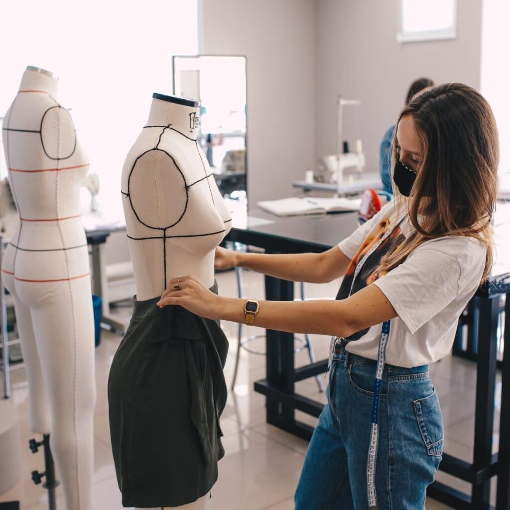 Fashion design student fitting skirt on dress form in a studio, wearing a mask, measuring tape around neck.