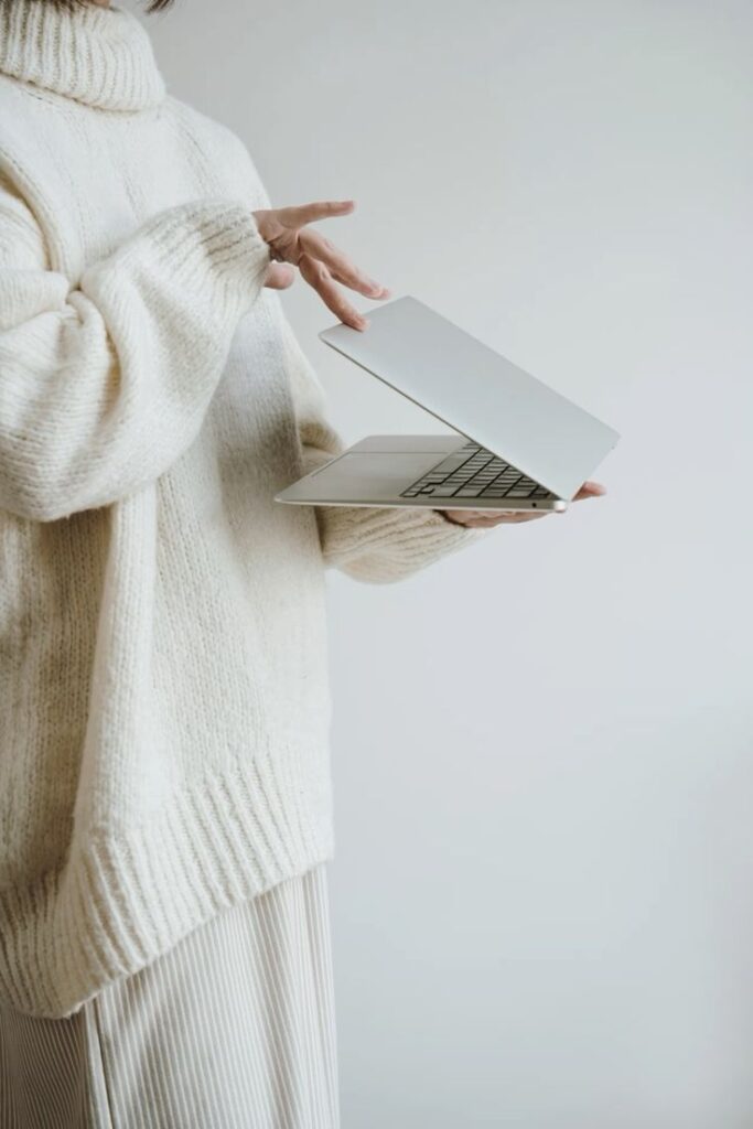 _ 4 Person in cozy sweater holding an open laptop, showcasing modern technology in a minimalistic style against a neutral background. | Sky Rye Design Person in cozy sweater holding an open laptop, showcasing modern technology in a minimalistic style against a neutral background.