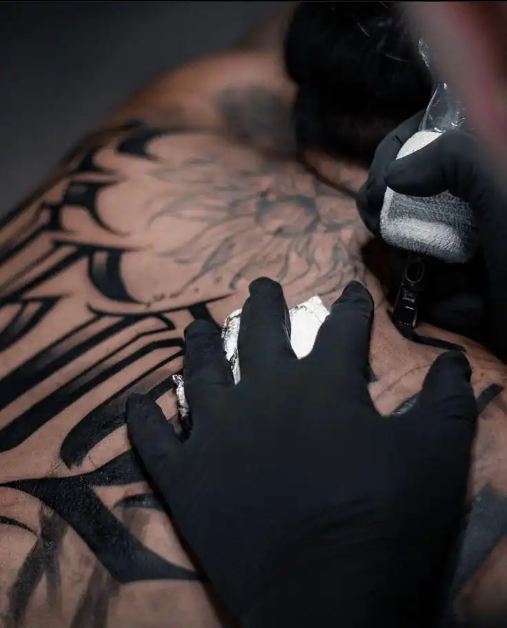 Tattoo artist with black gloves inking a complex design on a person's back. Close-up of tattoo creation in progress.