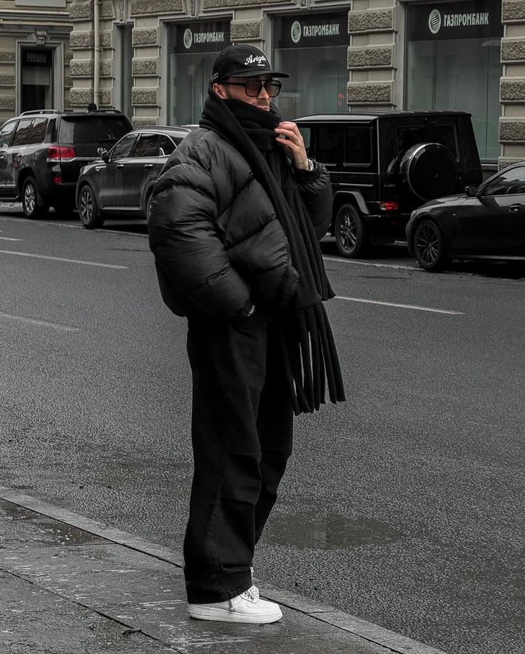Person in black puffer jacket, cap, scarf, and sneakers stands on urban street, with cars and a building in the background on a cloudy day.