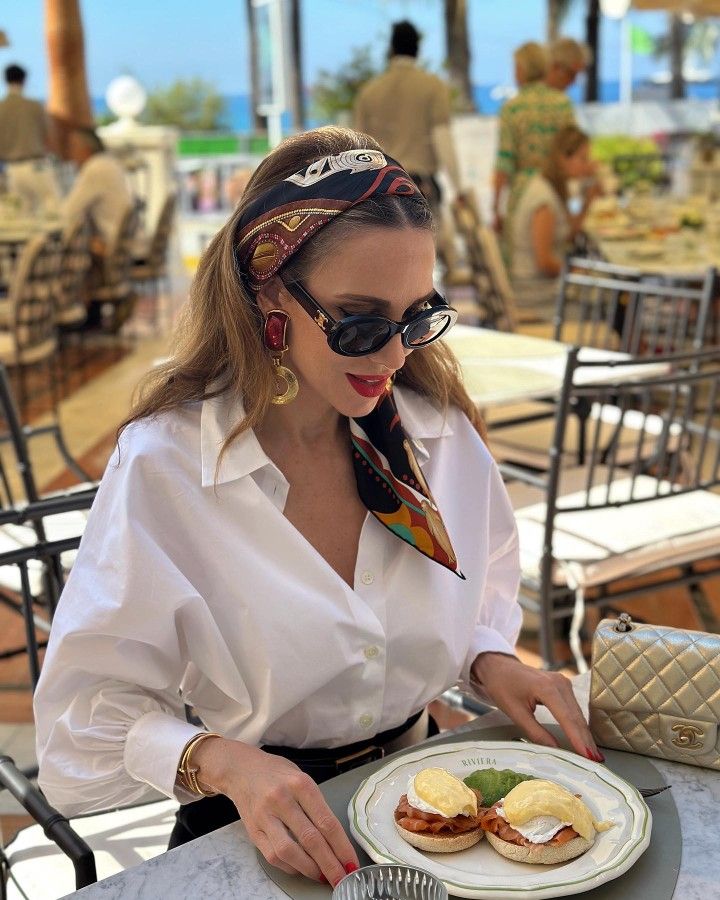 _ 40 Fashionable woman in sunglasses and scarf enjoys eggs benedict at outdoor café. Elegant brunch setting with ocean view in background. | Sky Rye Design Fashionable woman in sunglasses and scarf enjoys eggs benedict at outdoor café. Elegant brunch setting with ocean view in background.