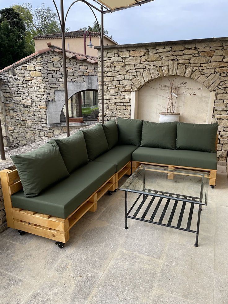 444d622bf5cc59604041d2766a368549 Elegant outdoor patio with green cushioned wooden pallet sofa, black metal glass coffee table, and rustic stone wall backdrop. | Sky Rye Design Elegant outdoor patio with green cushioned wooden pallet sofa, black metal glass coffee table, and rustic stone wall backdrop.
